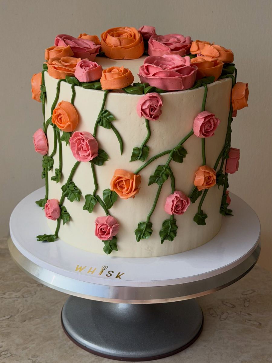 Torta blanca con rosas de buttercream en tonos coral y rosa que caen por los lados con hojas verdes